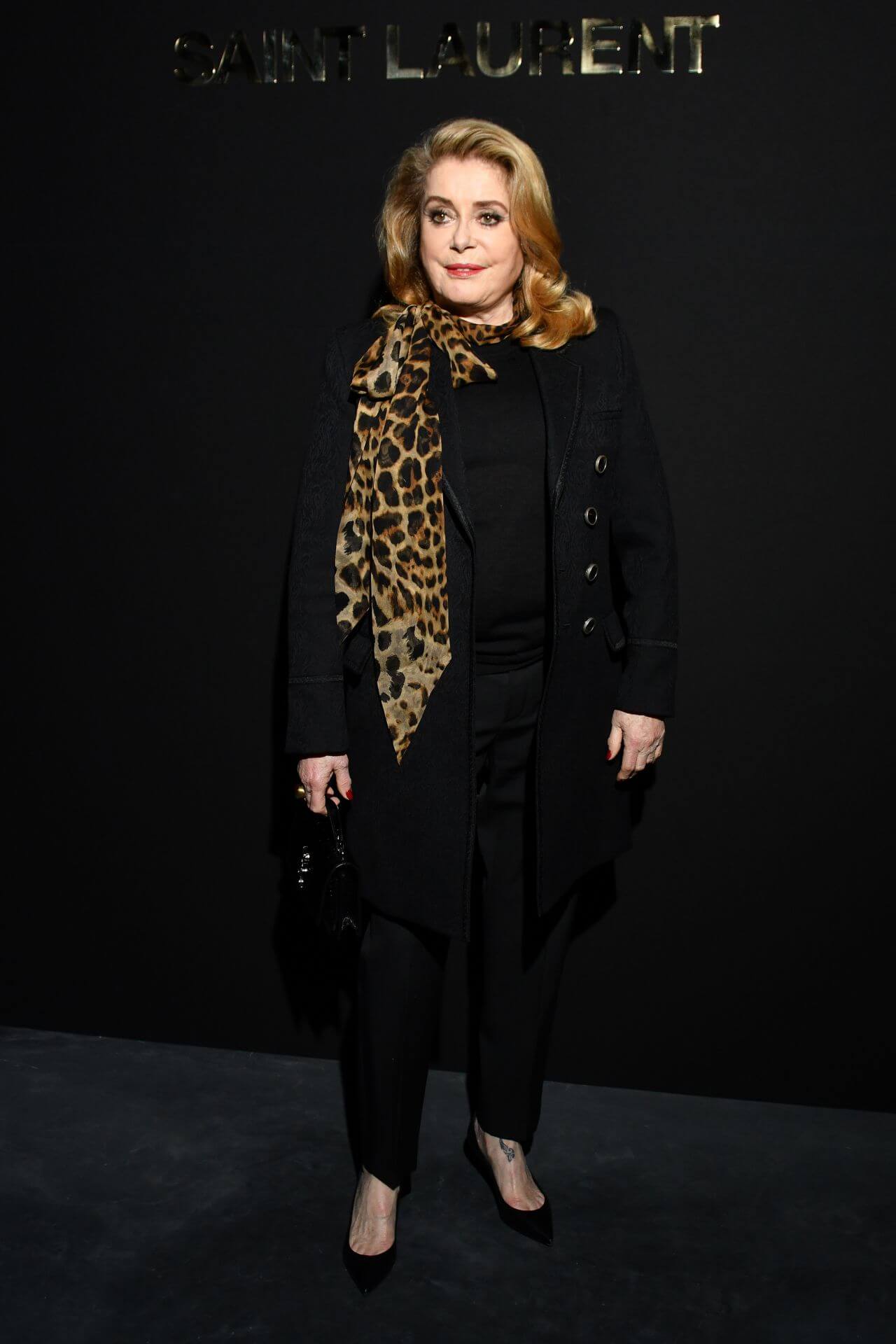 Catherine Deneuve In Black Long Coat Under Top & Pants With Tiger Print Neck scarf At Saint Laurent Fashion Show in Paris